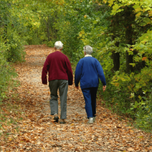 Older adult couple walking through a wooded area on a paved trail covered in fall leaves