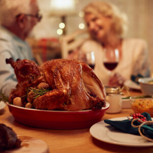 Delicious looking turkey on a platter on a table set for a family thanksgiving feast. Blurry background with a man & woman seated, conversing, & smiling while enjoying glasses of wine.